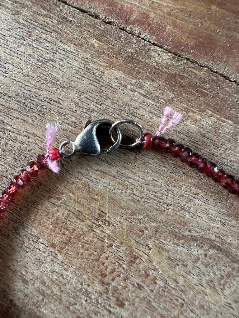 A close-up of a beaded bracelet with dark red beads, a silver lobster clasp, and two small pink tassels, laid on a rustic wooden surface.