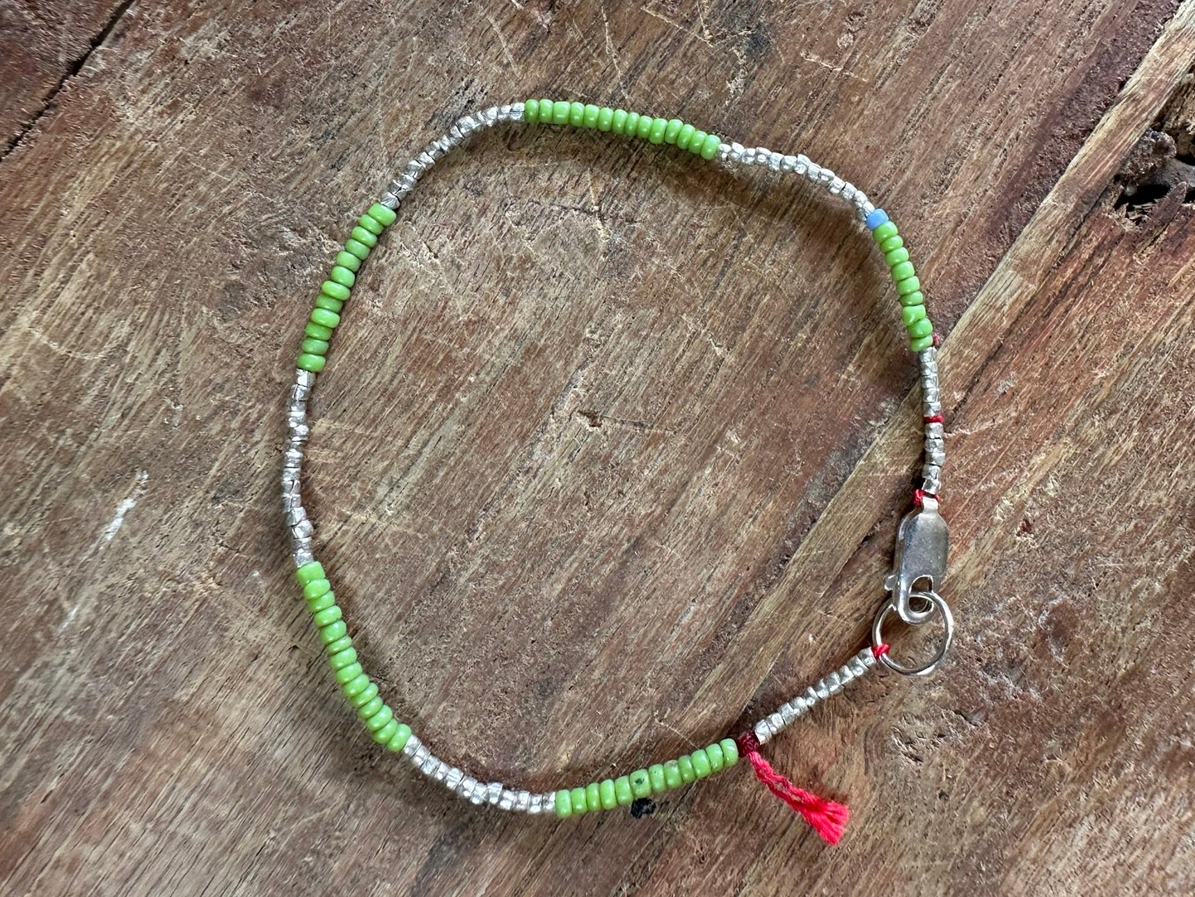 A beaded bracelet with lime green and silver beads, featuring a silver clasp and a small red tassel, displayed on a rustic wooden surface.