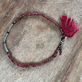 A beaded bracelet with dark red faceted beads, a small silver-toned spacer bead cluster, and a matching dark red tassel charm, displayed on a rustic wooden surface.