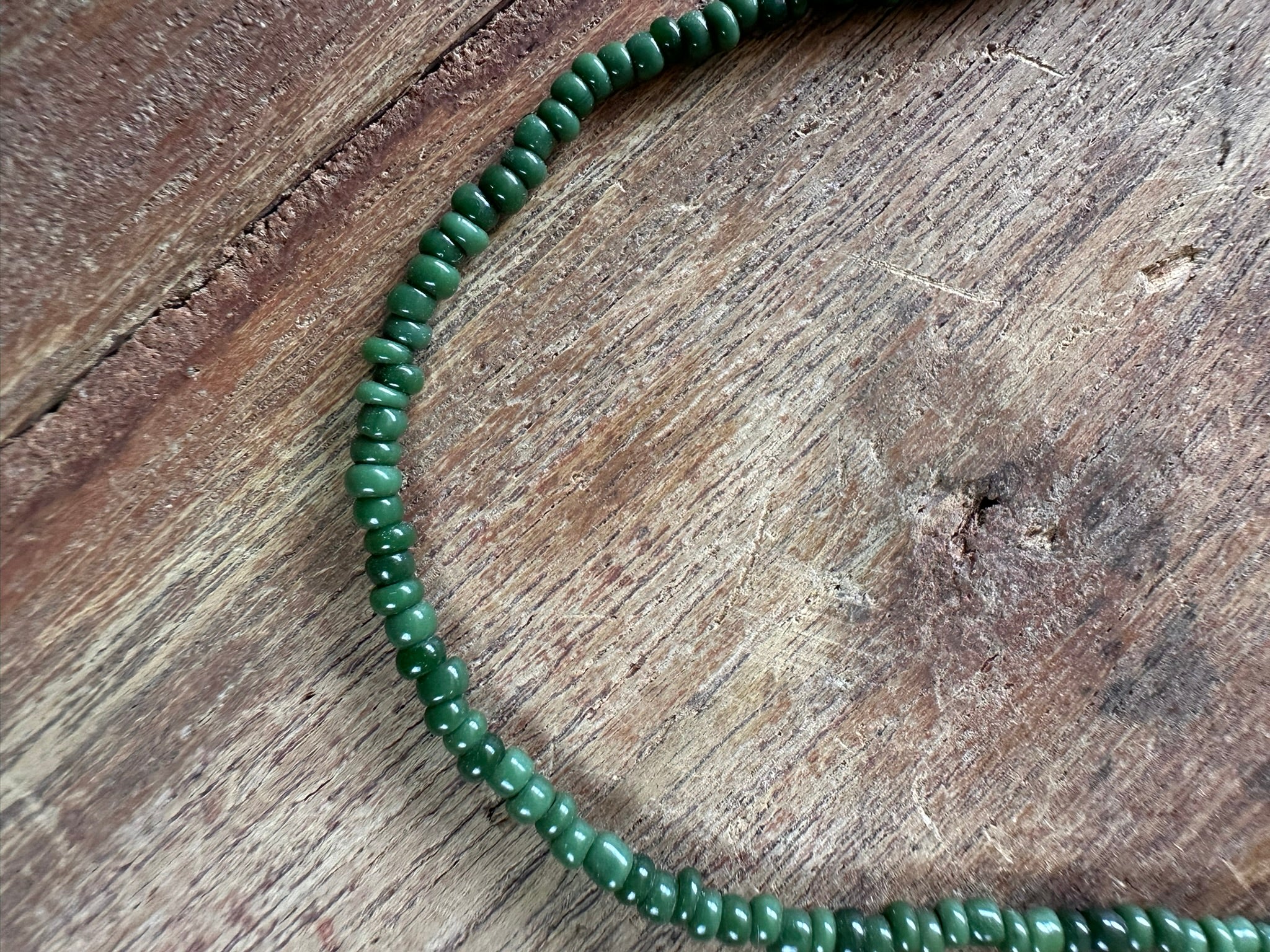 A close-up of a green beaded necklace or bracelet made of small, polished oval beads, arranged in a curve on a rustic, textured brown wooden surface.