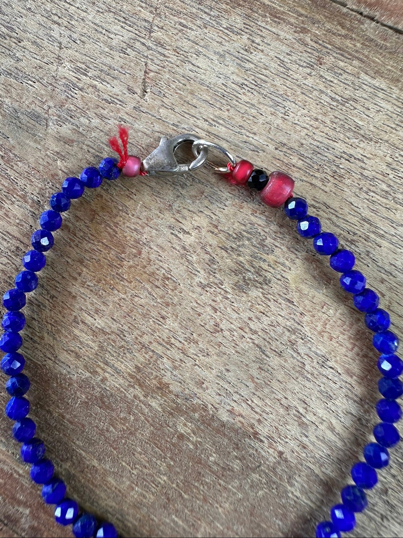 A beaded bracelet with faceted royal blue beads, a silver clasp, and small red and black accent beads, displayed on a rustic wooden surface.