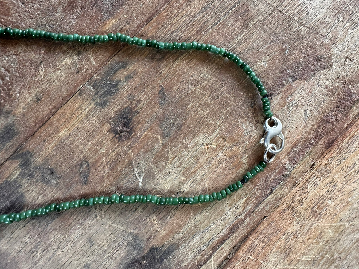 A close-up of a beaded necklace made of small dark green beads with a silver lobster clasp, laid on a rustic wooden surface.