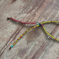 A handmade beaded necklace with a split design, featuring one strand of reddish-brown beads and another in yellow, red, and blue beads, laid out on a rustic wooden surface.