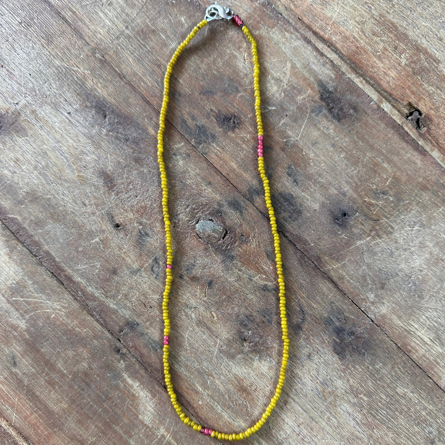 A beaded necklace made of small yellow beads with a few pink accent beads and a silver clasp, laid flat on a rustic wooden surface.