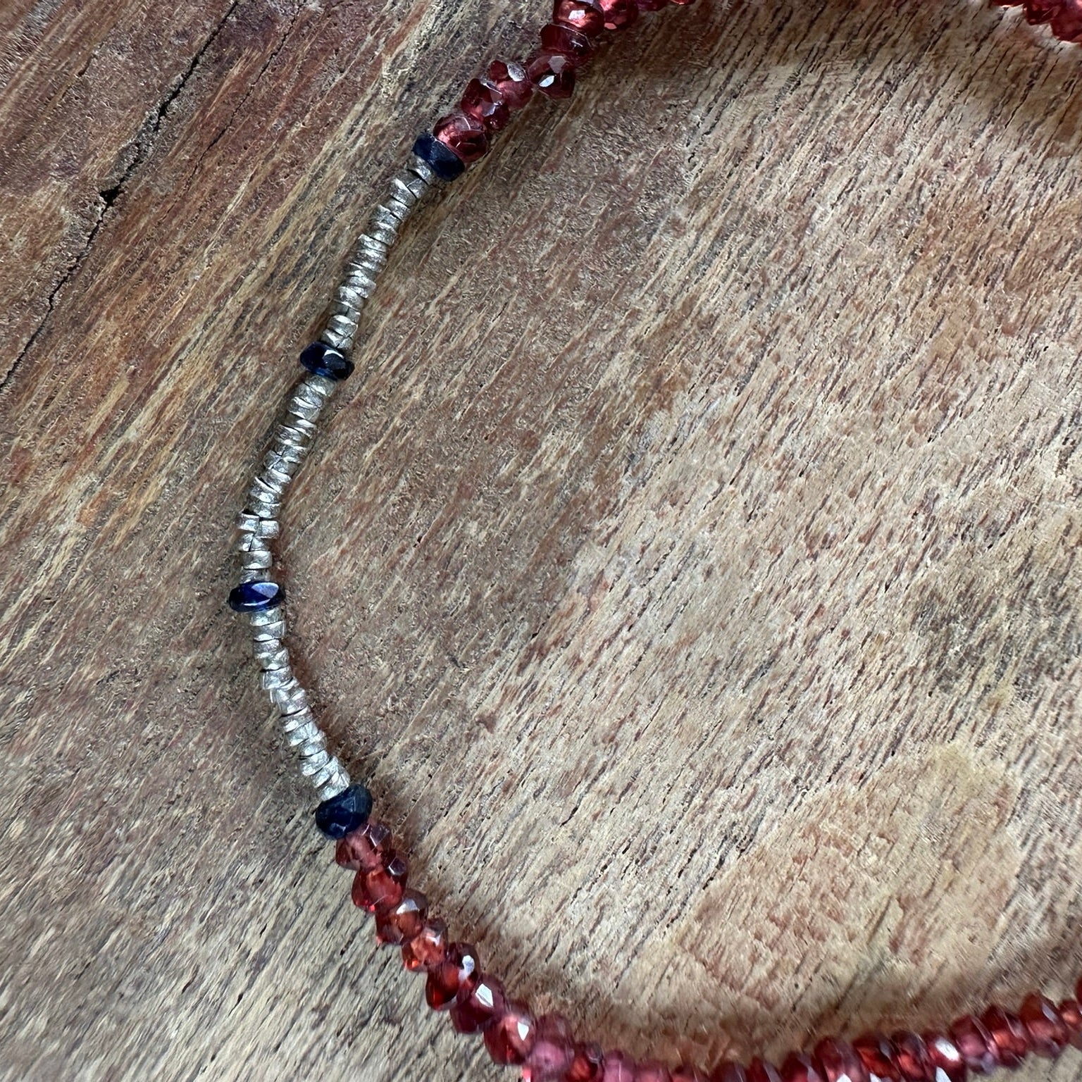 A close-up of a beaded necklace with faceted red beads, a textured silver section, and small dark blue beads, displayed on a rustic wooden surface.