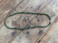 A beaded necklace made of small dark green beads with a silver-toned clasp, laid flat on a rustic wooden surface.