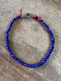 A beaded bracelet made of faceted deep blue beads, featuring a silver-toned lobster clasp, a small red bead, and a black bead near the clasp, displayed on a rustic wooden surface.