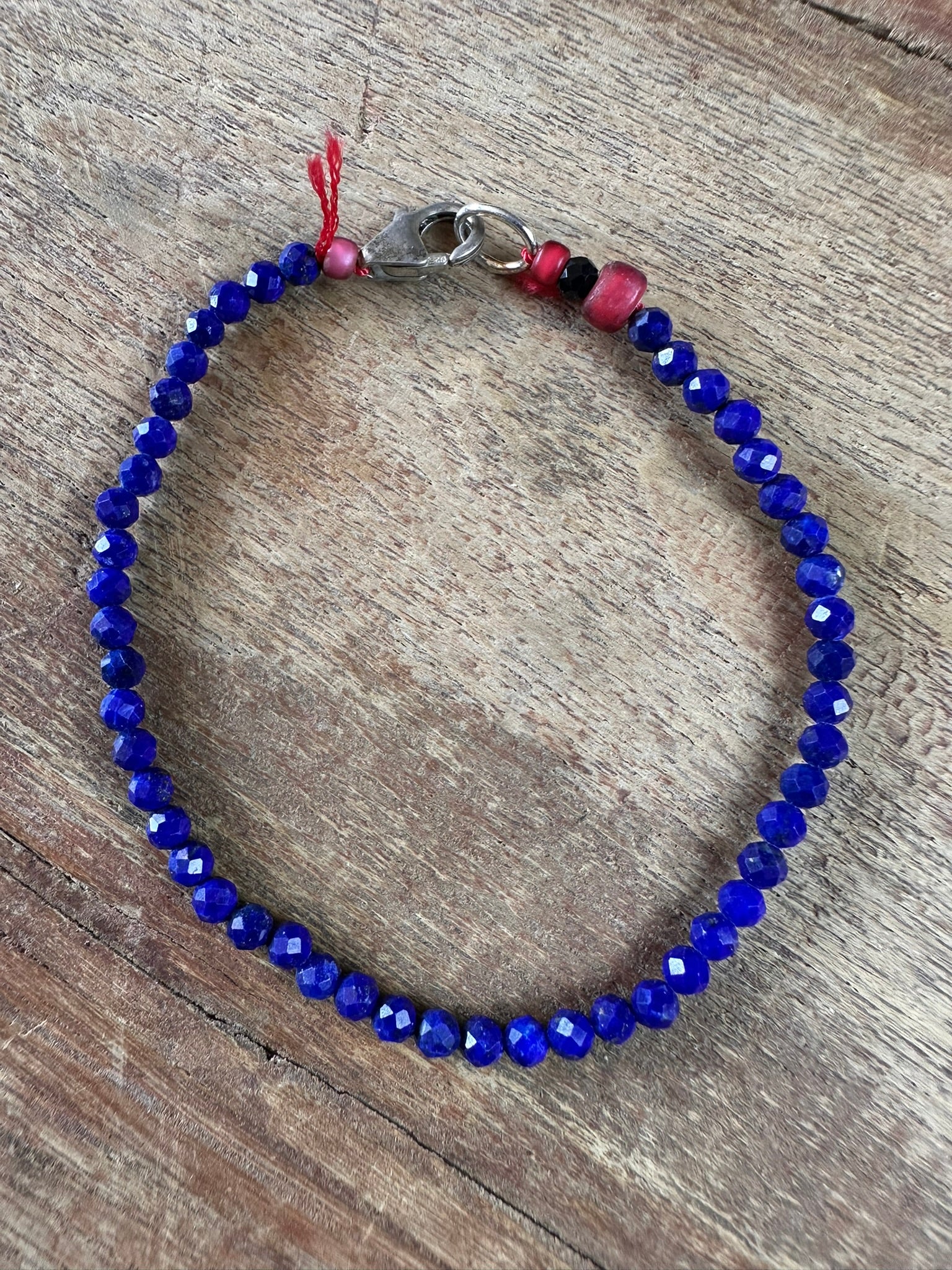 A beaded bracelet made of faceted deep blue beads, featuring a silver-toned lobster clasp, a small red bead, and a black bead near the clasp, displayed on a rustic wooden surface.
