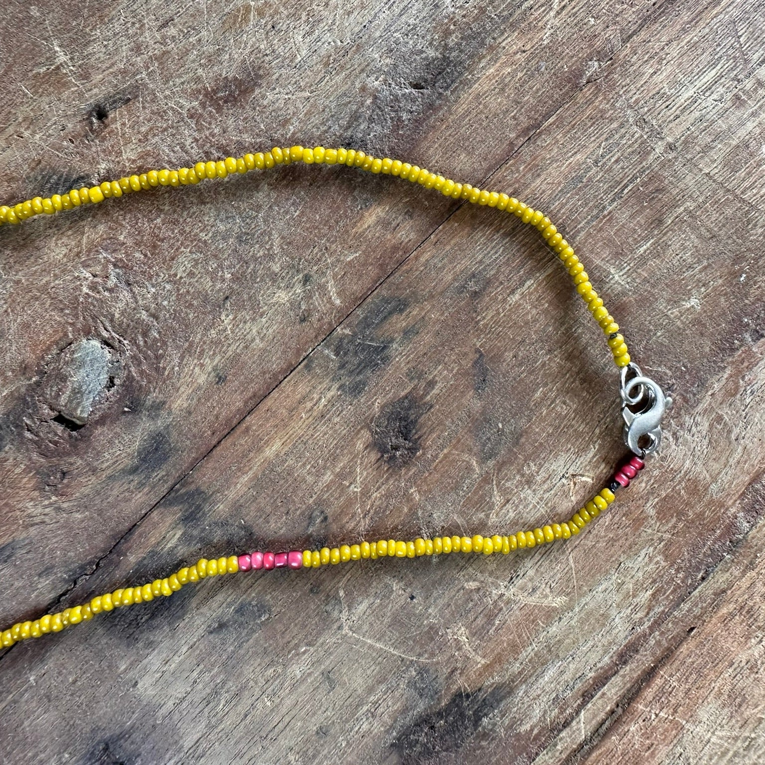 A beaded necklace made of small yellow beads with a few pink accent beads and a silver lobster clasp, laid on a rustic wooden surface.
