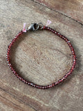 A beaded bracelet made of small faceted dark red beads with a silver lobster clasp, displayed on a rustic wooden surface.