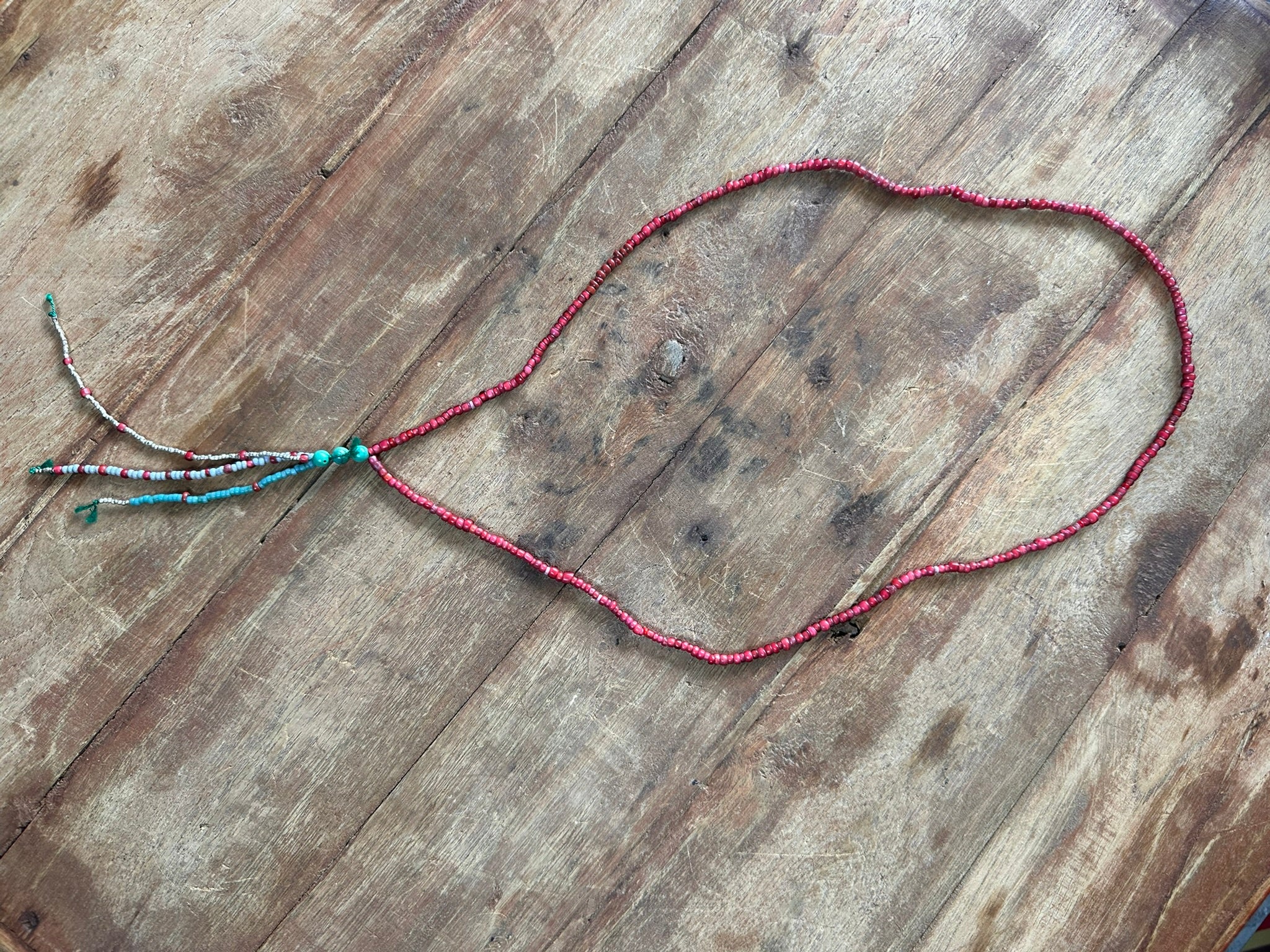 A beaded necklace with a long red strand and a short tassel detail featuring turquoise and white beads, laid flat on a rustic wooden surface.