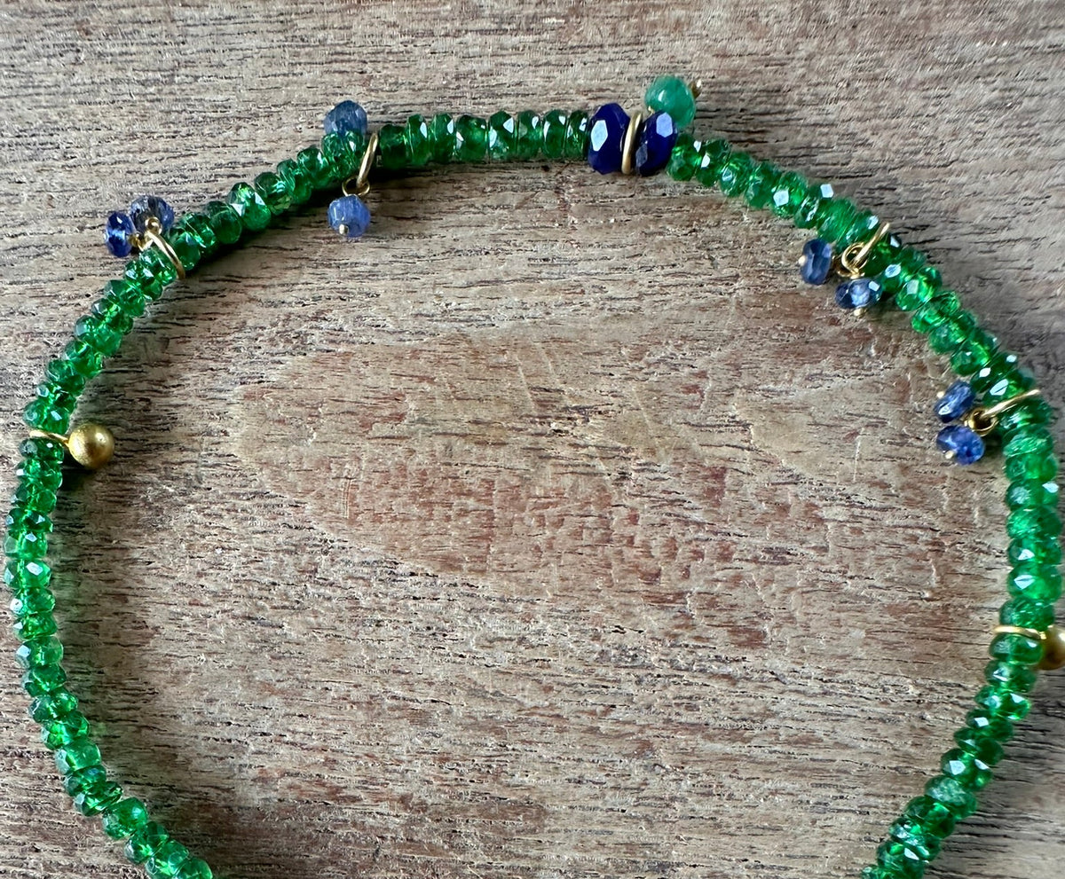 A beaded bracelet made of green faceted beads with small gold spacer beads, blue accent beads, and a gold clasp, displayed on a rustic wooden surface.