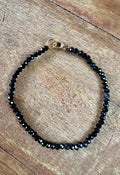 A beaded bracelet made of small faceted black beads with a gold clasp, displayed on a rustic wooden surface.