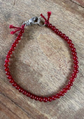A beaded bracelet made of small dark red beads with a silver-toned lobster clasp and red cord ties, displayed on a rustic wooden surface.