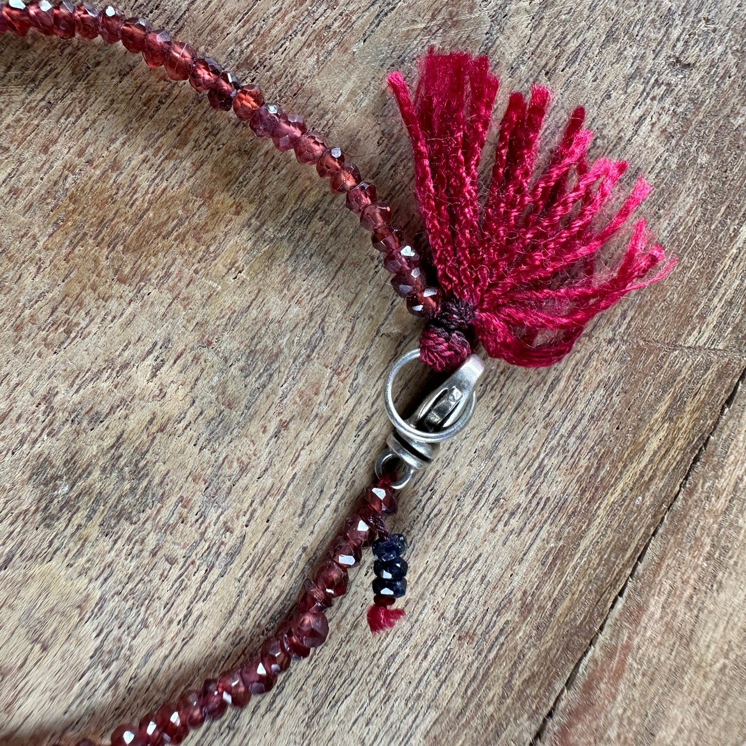 A close-up of a beaded necklace with deep red faceted beads, a matching red tassel, and a silver clasp, laid on a rustic wooden surface.