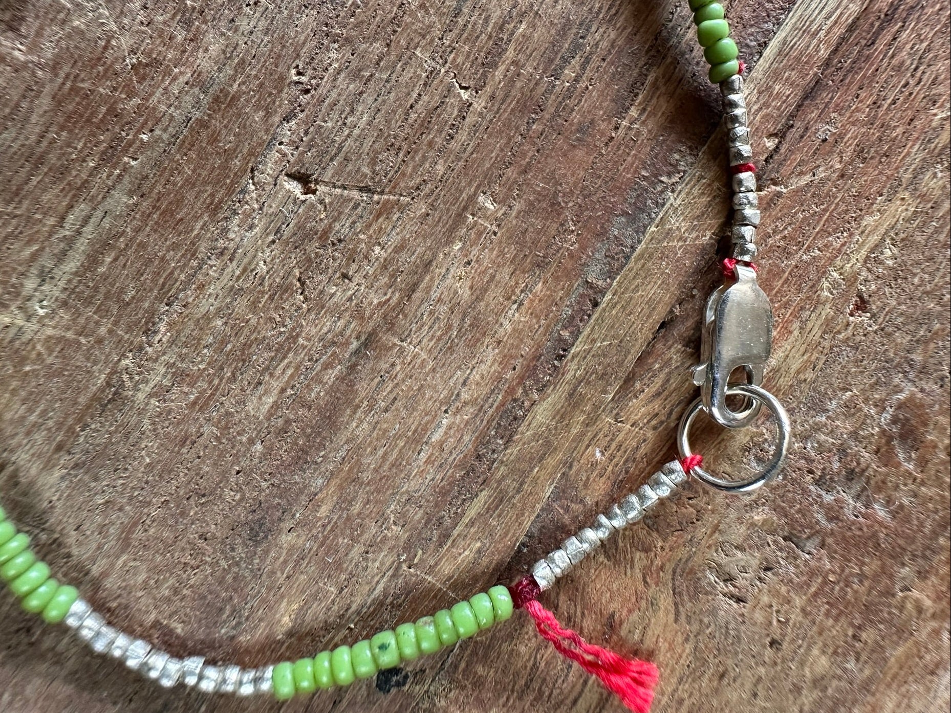 A beaded bracelet with light green and silver metallic beads, a silver clasp, and a small red tassel, displayed on a rustic wooden surface.
