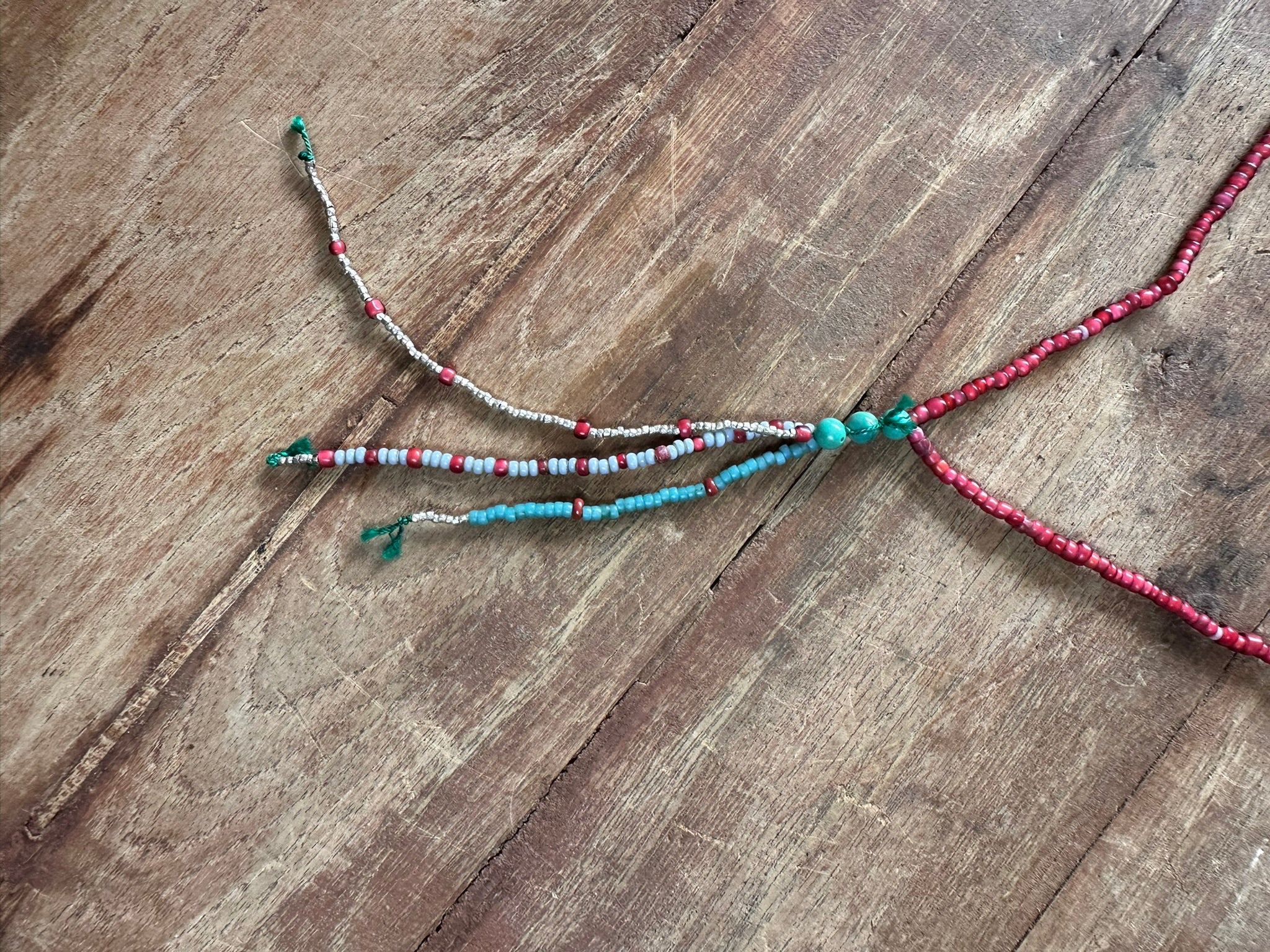 A handmade beaded necklace with three strands fanning out from a central knot, displayed on a rustic wooden surface. The strands feature red beads, light blue beads, white beads, and small turquoise-colored beads.