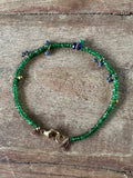 A beaded bracelet with green seed beads, small blue and gold spacer beads, and a gold clasp, displayed on a rustic wooden surface.