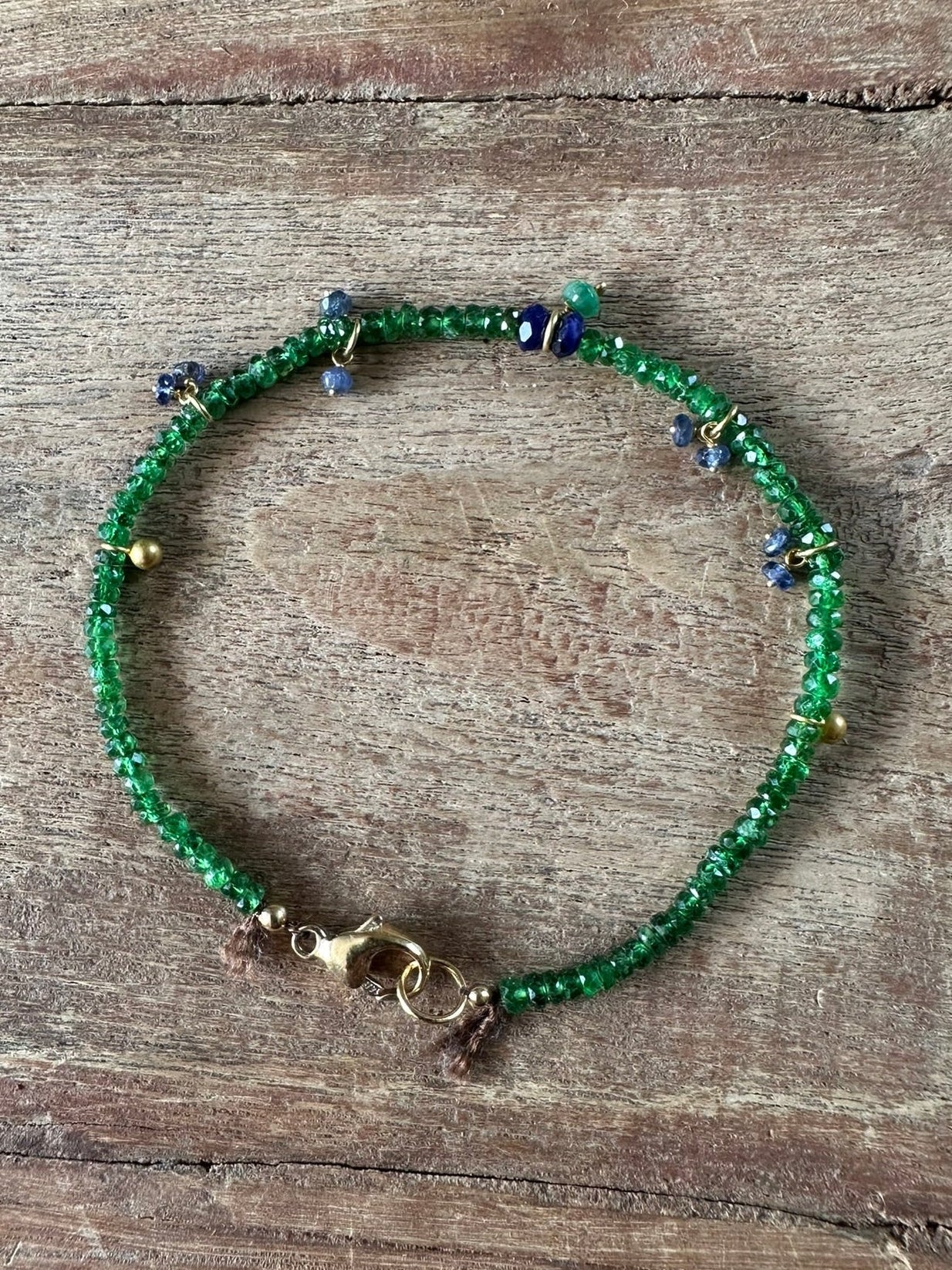A beaded bracelet with green seed beads, small blue and gold spacer beads, and a gold clasp, displayed on a rustic wooden surface.
