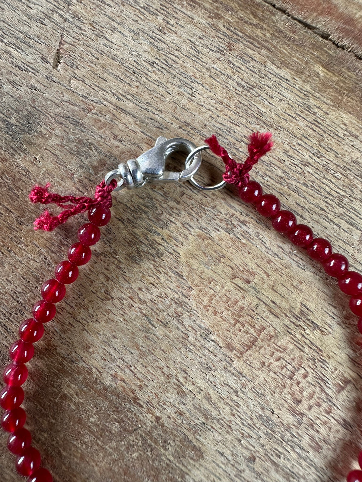A close-up of a red beaded bracelet with a silver lobster clasp and red cord ties, laid on a rustic wooden surface.