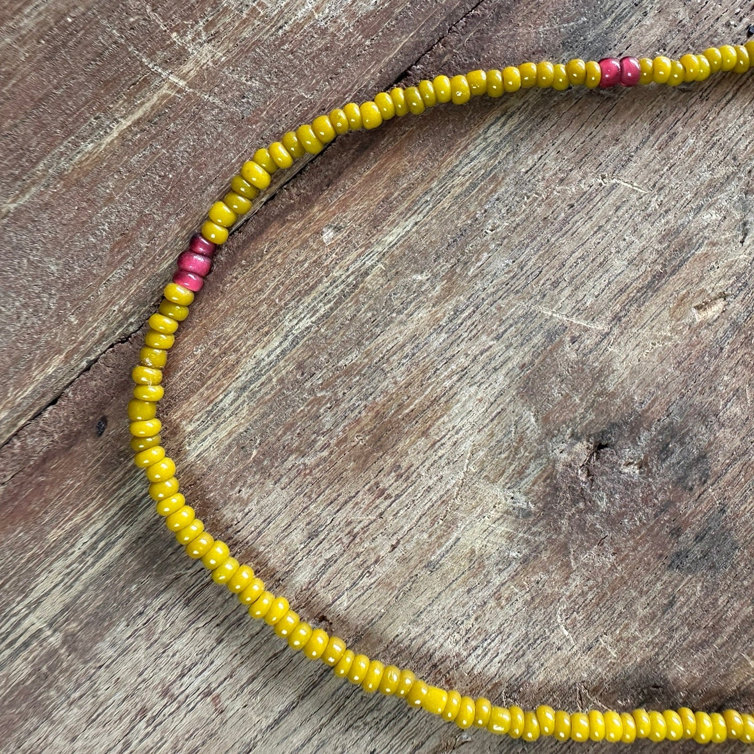 A beaded necklace made of small yellow beads with a few pink accent beads, laid out on a rustic wooden surface.