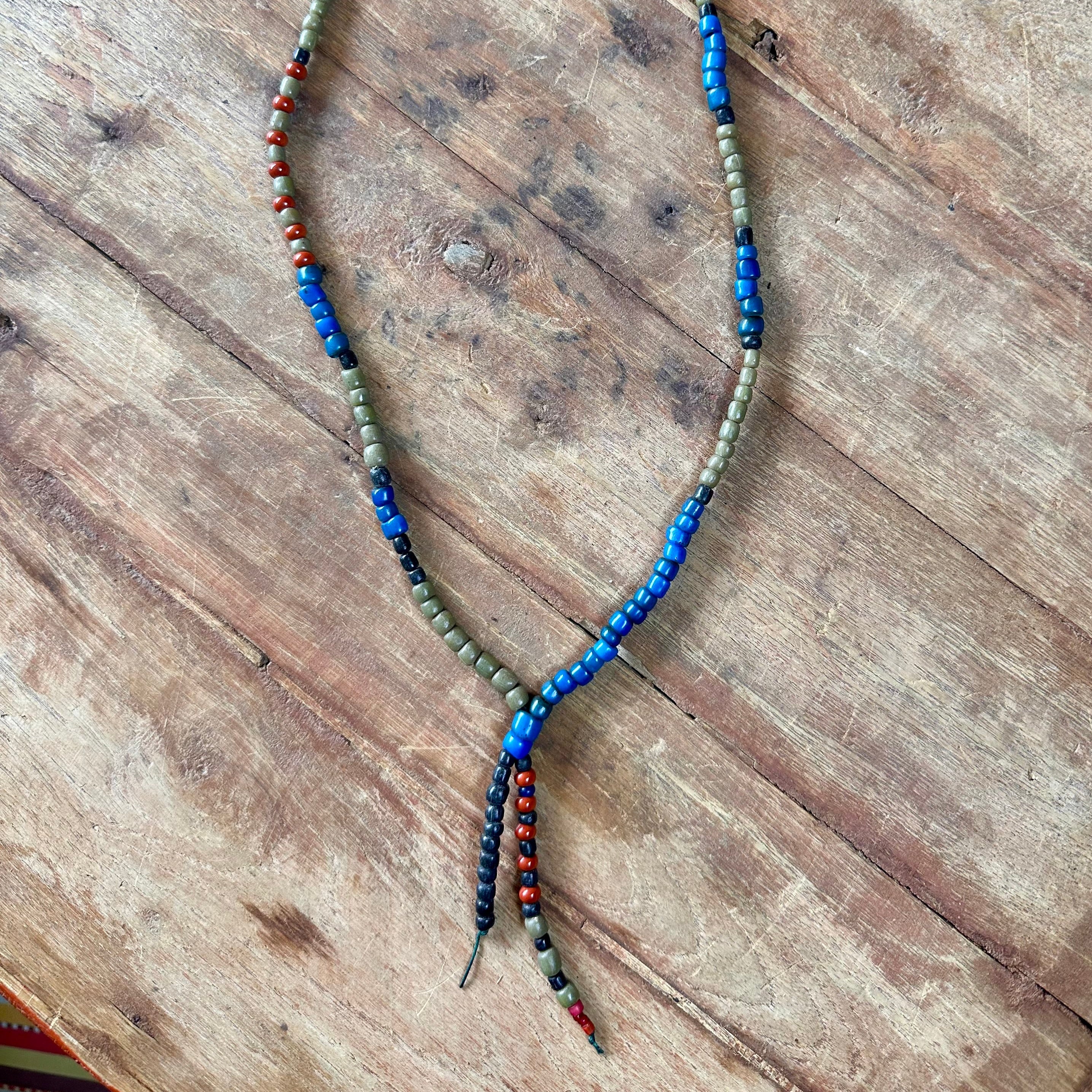 A beaded necklace with blue, olive green, and orange-red beads, laid flat on a rustic wooden surface.
