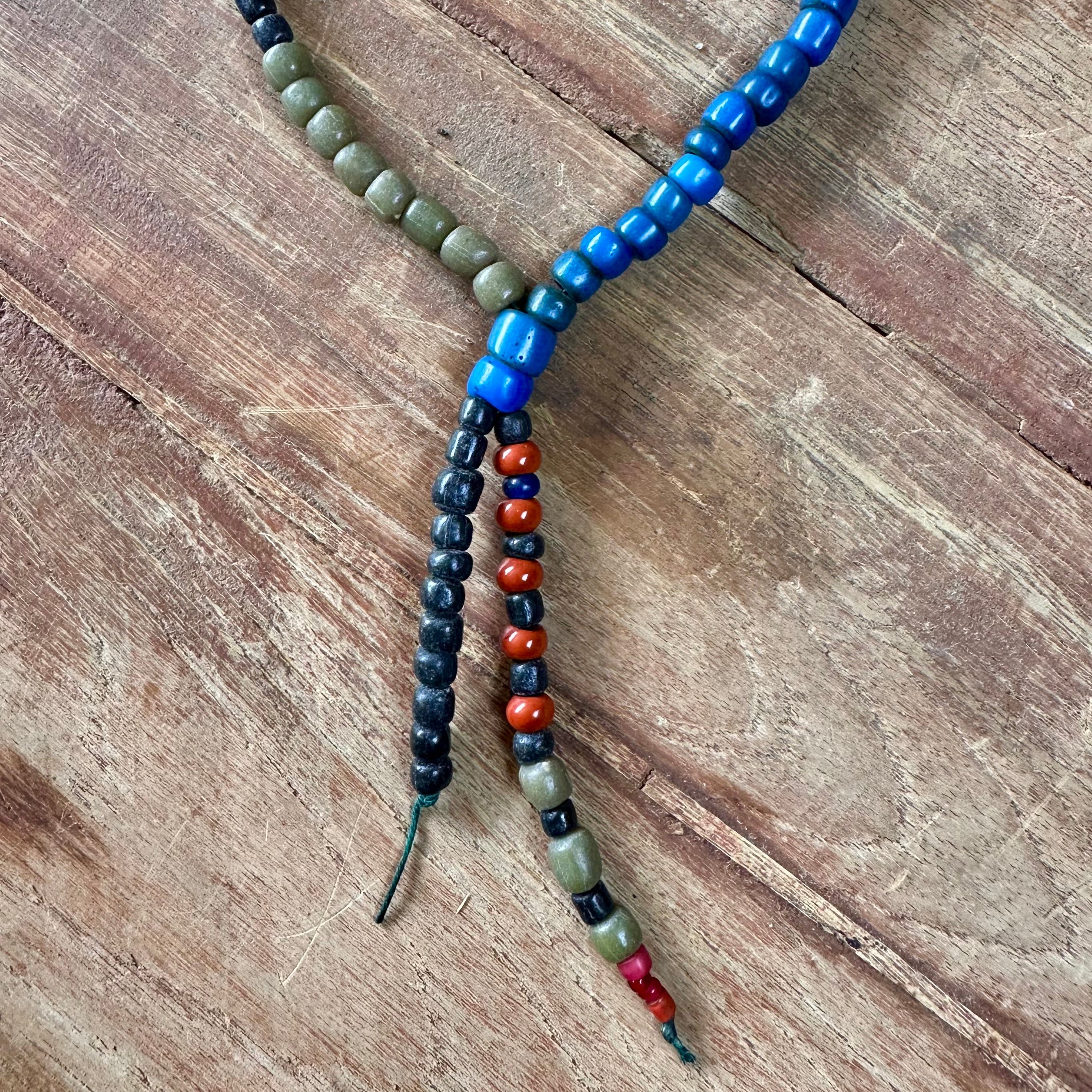 A handmade beaded necklace with a Y-shaped design, featuring olive green beads, bright blue beads, black beads, and red-orange beads, laid flat on a rustic wooden surface.