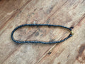 A beaded necklace made of small dark gray beads with a small gold-toned accent bead, laid out on a rustic wooden surface.