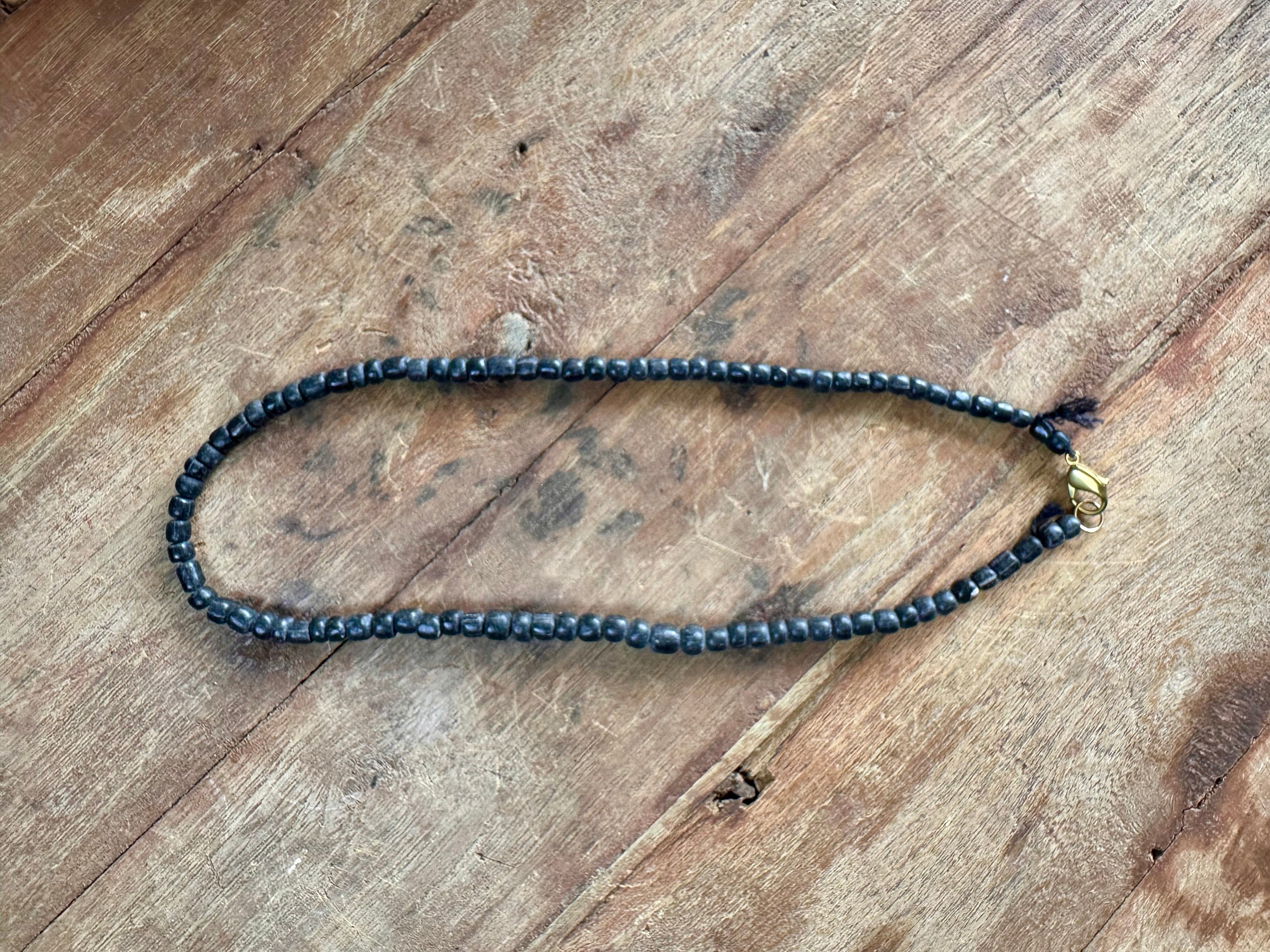 A beaded necklace made of small dark gray beads with a small gold-toned accent bead, laid out on a rustic wooden surface.