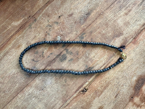 A beaded necklace made of small dark gray beads with a small gold-toned accent bead, laid out on a rustic wooden surface.