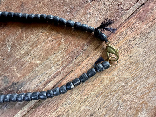 A close-up of a beaded necklace with dark, polished round beads and a brass clasp, laid on a rustic wooden surface.