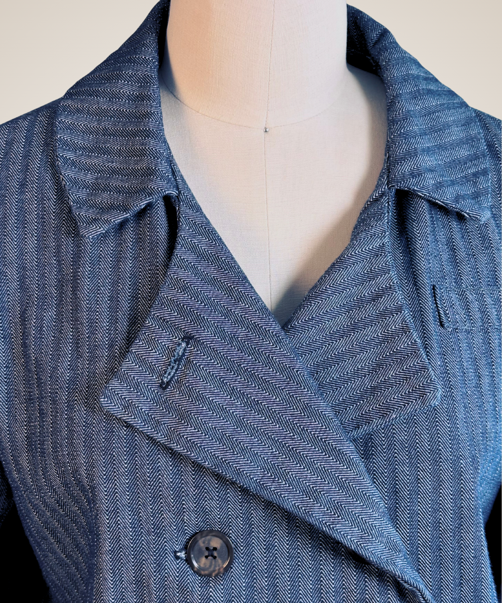 A close-up of a blue-gray textured herringbone jacket displayed on a cream-colored dress form mannequin. The jacket features a wide collar, a notched lapel, and a single visible dark button.