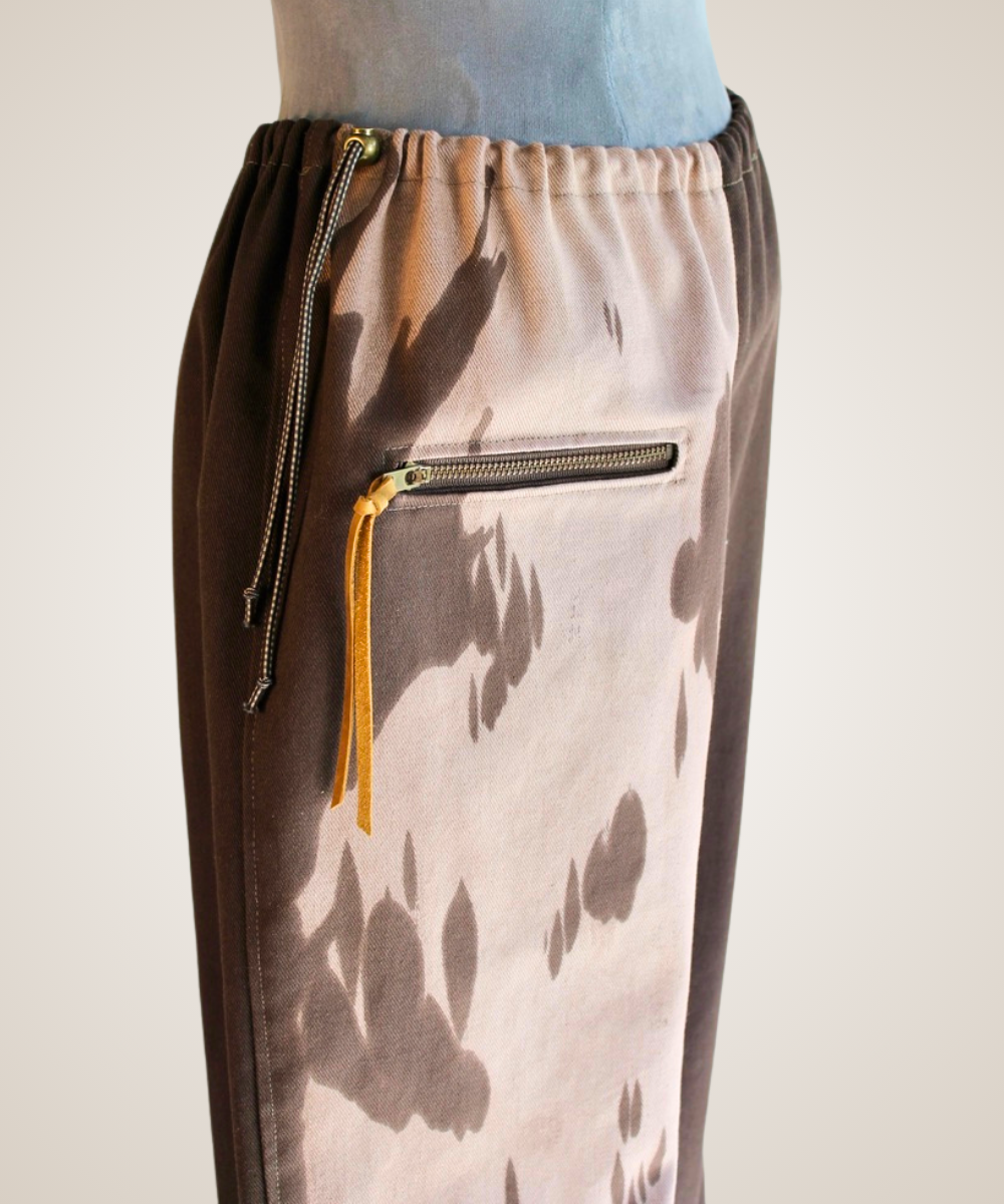 A close-up of a pair of wide-leg pants displayed on a mannequin torso. The pants feature a split design: one side has a brown leather patch pocket and a brown-and-beige abstract print, while the other side is a solid dark brown suede-like fabric.