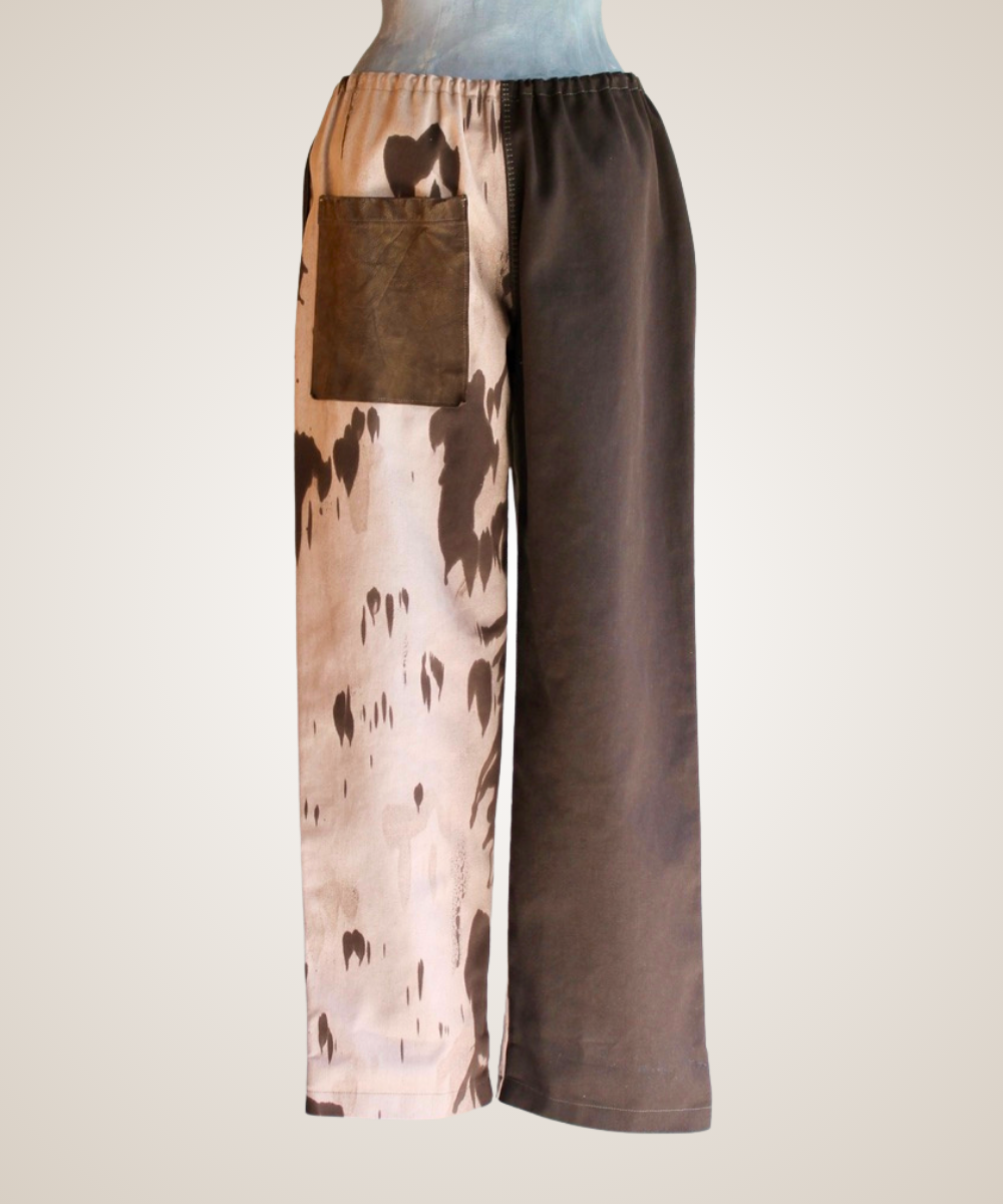 Wide-leg pants displayed on a mannequin torso against a plain light background. The pants have an asymmetrical split design: one side is light beige with dark brown abstract spots, and the other side is solid dark brown.