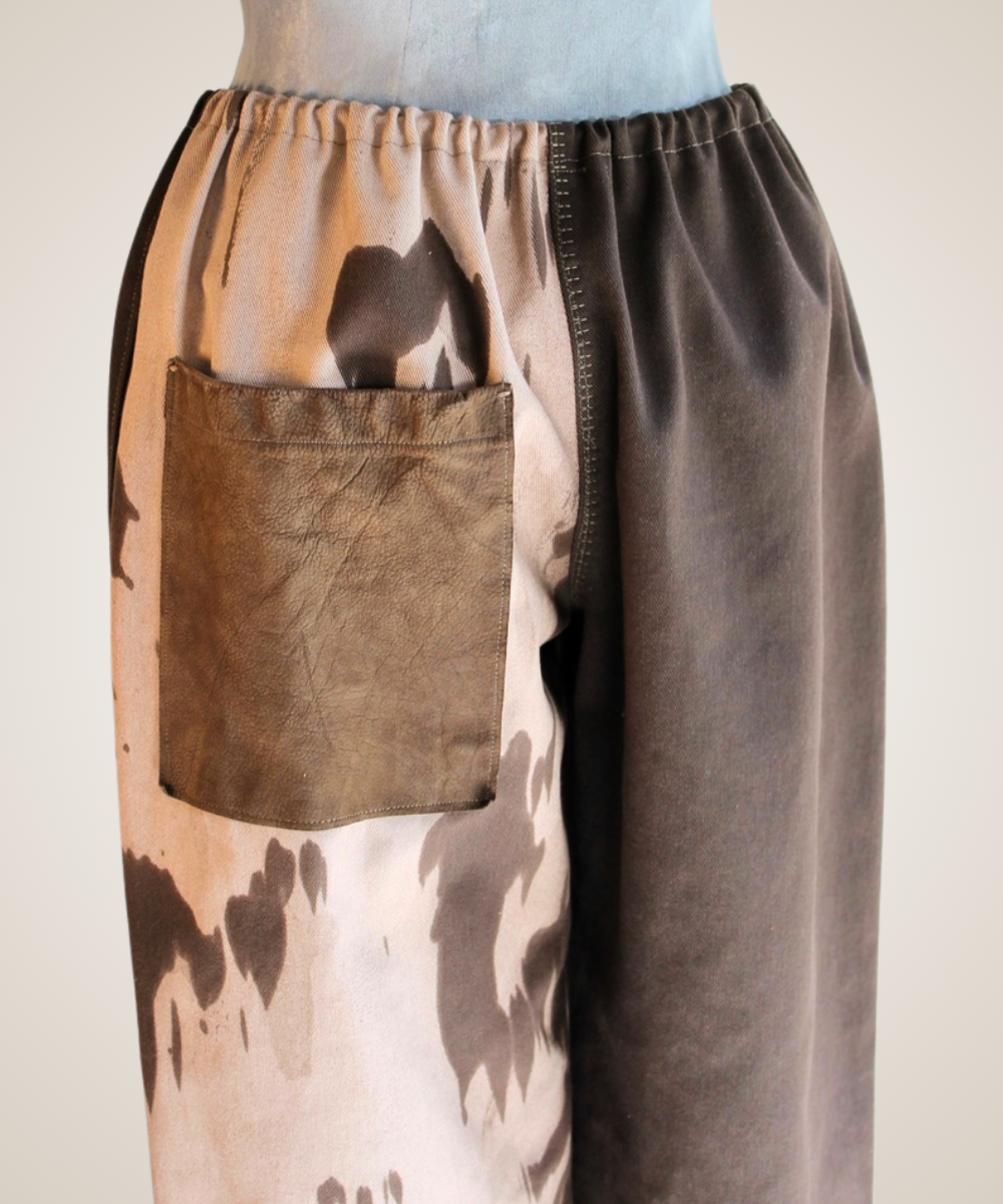 A close-up of a pair of wide-leg pants displayed on a mannequin torso. The pants feature a split design: one side has a brown leather patch pocket and a brown-and-beige abstract print, while the other side is a solid dark brown suede-like fabric.