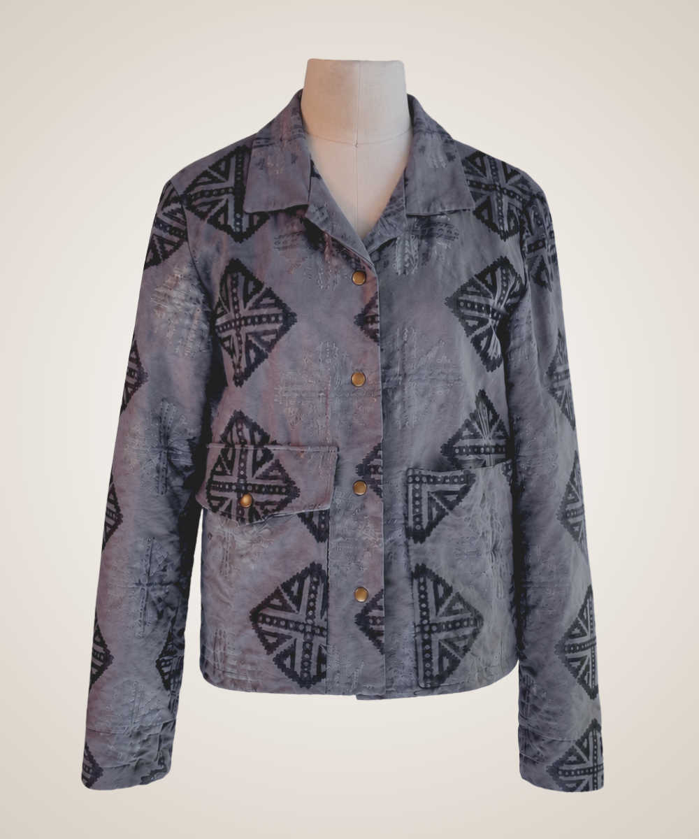 A long-sleeve button-front jacket displayed on a mannequin torso against a plain light beige background. The jacket has a muted gray-purple base with a dark geometric tribal-style pattern and small brown buttons.