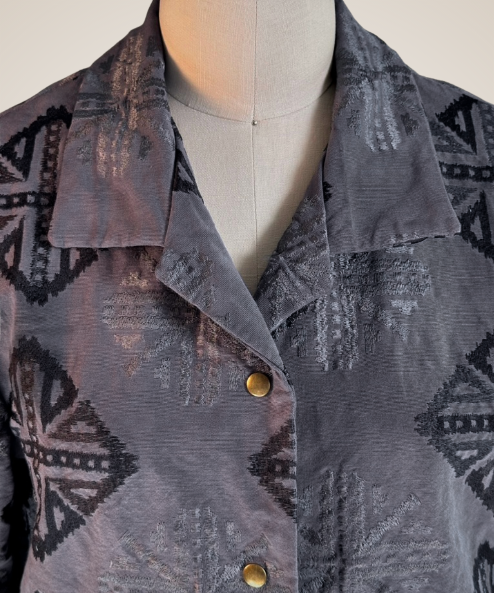 A close-up of a gray patterned button-up shirt jacket displayed on a dress form mannequin. The fabric features a textured, woven geometric tribal pattern in shades of gray and black, with three gold-tone buttons down the front.