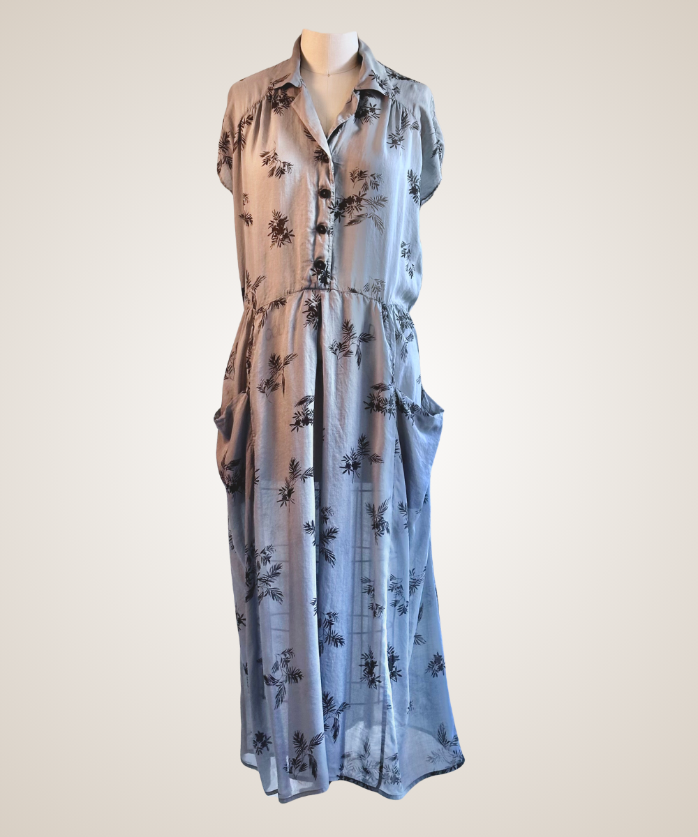 A sleeveless light blue midi dress with a black palm tree print, featuring a button-up front and side pockets, displayed on a mannequin against a plain beige background.