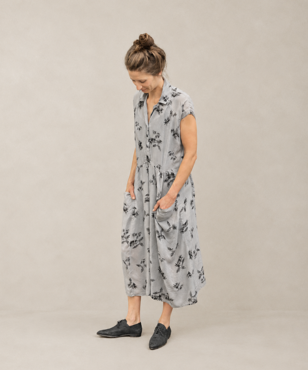 A woman wearing a light gray sleeveless midi dress with a black floral print, standing against a neutral beige studio background.