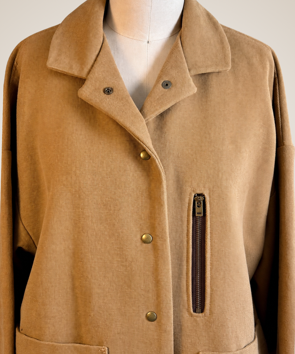 A tan wool-blend jacket with a notched lapel collar, brass-tone snap buttons, and a vertical zippered pocket on the left side, displayed on a mannequin torso.