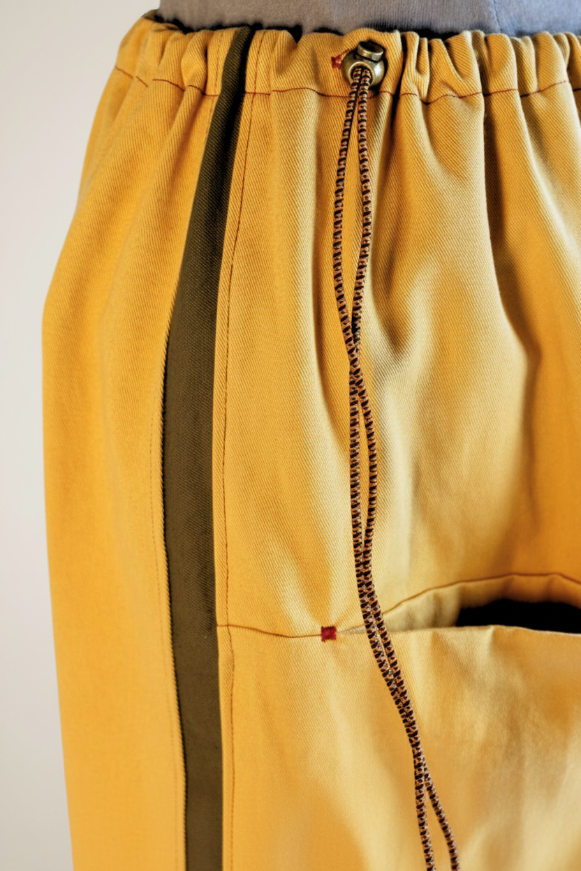 Close-up of mustard yellow pants with a dark olive vertical stripe, a dark brown drawstring, and a side pocket.